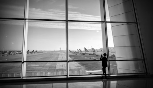 A black and white photograph of an airport runway viewed through large glass panels. Several airplanes are parked in the distance, and a solitary figure stands by the window, possibly taking a photograph.