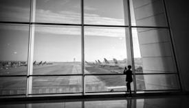 A black and white photograph of an airport runway viewed through large glass panels. Several airplanes are parked in the distance, and a solitary figure stands by the window, possibly taking a photograph.