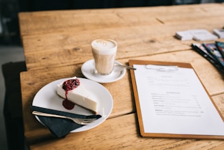 A cozy cafe scene featuring a slice of cheesecake garnished with berries on a white plate, accompanied by a black napkin and a fork. A glass of latte with a frothy top is placed beside the cake, and a menu on a clipboard lies nearby on the rustic wooden table.