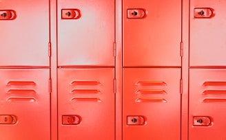 A row of vibrant red metal lockers with visible handles and keyholes. Each locker has a smooth surface with vents and distinctive lines, arranged in a uniform pattern.
