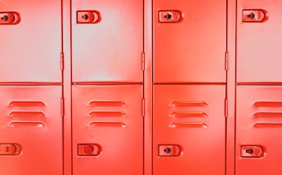 A row of vibrant red metal lockers with visible handles and keyholes. Each locker has a smooth surface with vents and distinctive lines, arranged in a uniform pattern.