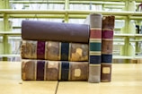 A stack of old, worn books with leather bindings rests on a wooden table. The books have visible signs of wear, including frayed edges and fading on the spines. They are positioned in front of empty library shelves that are yellowish in color.