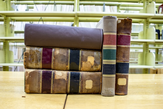 A stack of old, worn books with leather bindings rests on a wooden table. The books have visible signs of wear, including frayed edges and fading on the spines. They are positioned in front of empty library shelves that are yellowish in color.
