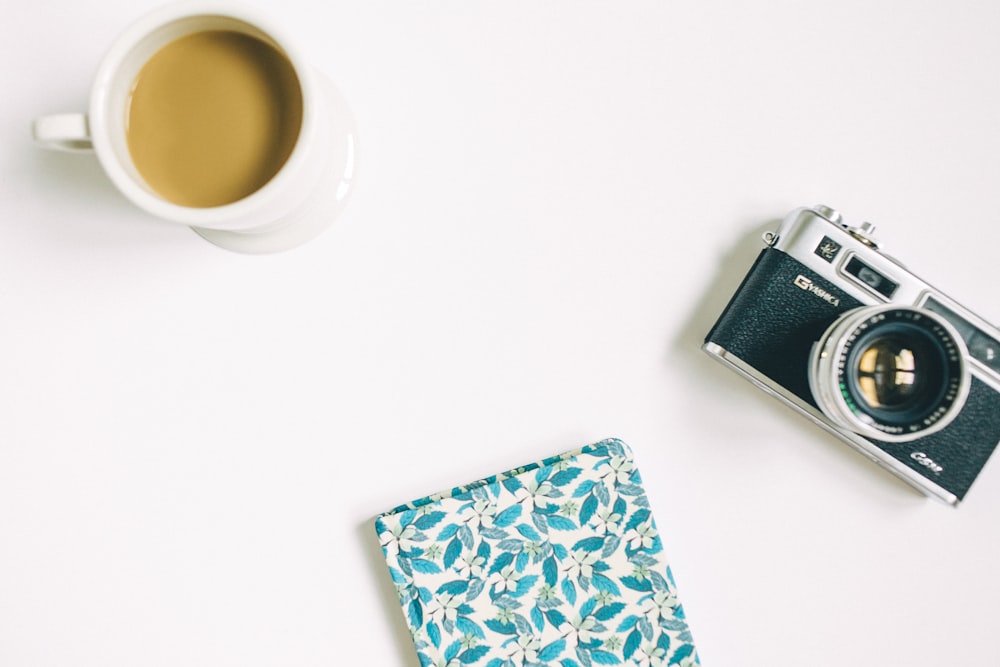 black and grey SLR camera, floral notebook, and white ceramic mug