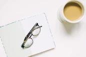 A stylish pair of eyeglasses resting on an open book beside a cup of coffee.