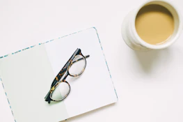 A stylish pair of eyeglasses resting on an open book beside a cup of coffee.