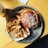 A metal tray holds a fresh burger topped with grated cheese, accompanied by a side of crispy, golden fries also sprinkled with cheese. A drink sits nearby on a wooden table with a receipt or order ticket partially visible in the background.