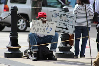 A person is sitting on the sidewalk holding a sign that reads 'I'm just hungry'. They are wearing a red cap and jeans. Around them are street posts and a sign instructing pedestrians to cross using the other side. The background includes a parked vehicle and another person standing nearby.