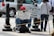 A person is sitting on the sidewalk holding a sign that reads 'I'm just hungry'. They are wearing a red cap and jeans. Around them are street posts and a sign instructing pedestrians to cross using the other side. The background includes a parked vehicle and another person standing nearby.