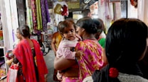 A bustling market scene with several people engaging in shopping. A woman in a bright pink and yellow saree carries a child, who is looking toward the camera. Colorful textiles and garments hang around the shop, creating a vibrant atmosphere. Other individuals are partially visible, focused on their activities.