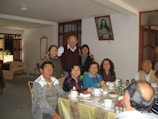 A group of people of various ages are gathered around a dining table indoors, enjoying a meal together. The table is covered with a light-colored tablecloth and set with cups, saucers, utensils, and various boxes. The room is decorated in a homey style, with a framed picture of Jesus on the wall. Several of the individuals are smiling, suggesting a warm, familial atmosphere.