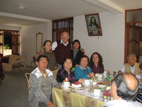 A group of people of various ages are gathered around a dining table indoors, enjoying a meal together. The table is covered with a light-colored tablecloth and set with cups, saucers, utensils, and various boxes. The room is decorated in a homey style, with a framed picture of Jesus on the wall. Several of the individuals are smiling, suggesting a warm, familial atmosphere.