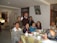 A group of people of various ages are gathered around a dining table indoors, enjoying a meal together. The table is covered with a light-colored tablecloth and set with cups, saucers, utensils, and various boxes. The room is decorated in a homey style, with a framed picture of Jesus on the wall. Several of the individuals are smiling, suggesting a warm, familial atmosphere.