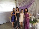 Four women are standing together, posing for a photo at what appears to be an event or celebration. They are dressed in formal attire, with elegant dresses in various colors. Behind them is a backdrop of draped white and purple fabric with tables adorned with floral arrangements and a decorative cake.