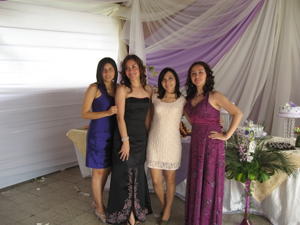 Four women are standing together, posing for a photo at what appears to be an event or celebration. They are dressed in formal attire, with elegant dresses in various colors. Behind them is a backdrop of draped white and purple fabric with tables adorned with floral arrangements and a decorative cake.