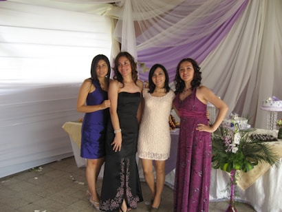 Four women are standing together, posing for a photo at what appears to be an event or celebration. They are dressed in formal attire, with elegant dresses in various colors. Behind them is a backdrop of draped white and purple fabric with tables adorned with floral arrangements and a decorative cake.