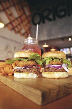 Two gourmet burgers topped with onion rings, fresh lettuce, and red onion slices are placed on a wooden board. Waffle fries are positioned to the side. In the background, a mason jar glass filled with a pink beverage and a straw is visible. The setting includes ambient lighting with a rustic and casual decor theme.