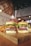 Two gourmet burgers topped with onion rings, fresh lettuce, and red onion slices are placed on a wooden board. Waffle fries are positioned to the side. In the background, a mason jar glass filled with a pink beverage and a straw is visible. The setting includes ambient lighting with a rustic and casual decor theme.