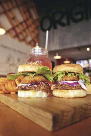 Two gourmet burgers topped with onion rings, fresh lettuce, and red onion slices are placed on a wooden board. Waffle fries are positioned to the side. In the background, a mason jar glass filled with a pink beverage and a straw is visible. The setting includes ambient lighting with a rustic and casual decor theme.