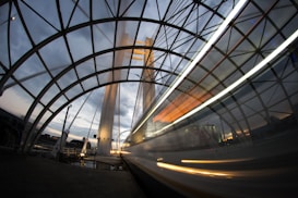A dynamic urban scene captures a large structure with an architectural arch design, likely a bridge or tunnel. The image is taken with a long exposure, resulting in a blurred trail of light indicating fast-moving traffic, possibly a train or car, passing through. The sky above is a mix of gray and blue, suggesting either dawn or dusk. The infrastructure is illuminated with lights, adding a dramatic contrast to the scene.