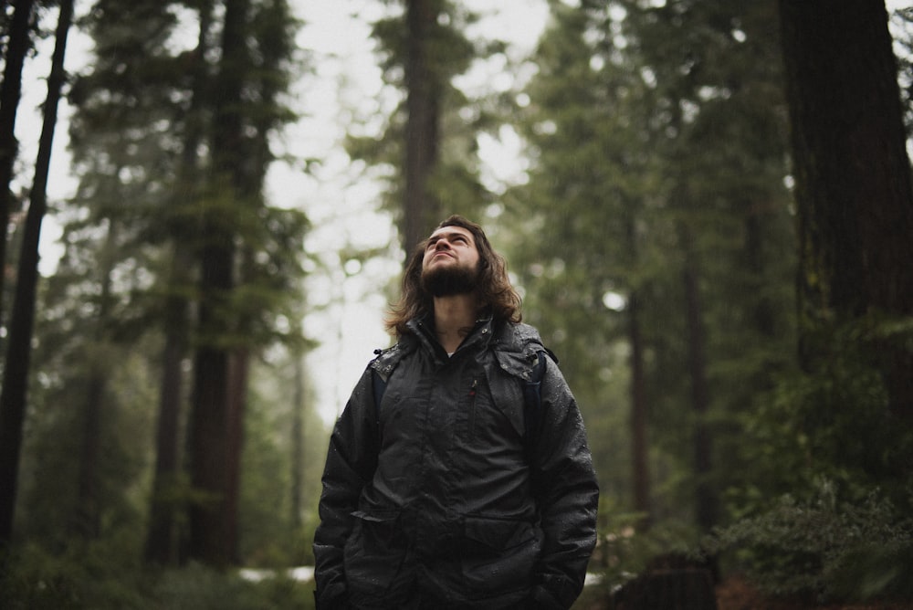 man wearing black jacket looking up surrounded with tall trees