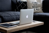 A sleek laptop with an illuminated logo is placed on a wooden coffee table. The table also holds a tablet and a smartphone. In the background, there's a dark gray sofa and a decorative pillow with text.