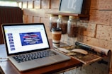 A cozy corner in a café with a laptop displaying a presentation on HTML, coffee jars mounted on the wall, a takeaway coffee cup, a wireless mouse, and an open magazine on the table. The setting suggests a warm and inviting atmosphere for work or relaxation.