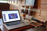 A cozy corner in a café with a laptop displaying a presentation on HTML, coffee jars mounted on the wall, a takeaway coffee cup, a wireless mouse, and an open magazine on the table. The setting suggests a warm and inviting atmosphere for work or relaxation.