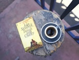 A weathered round wooden table holds an old paperback book, 'Travels with Charley' by John Steinbeck, alongside a small cup of espresso on a saucer. The table appears rustic with visible signs of age, adding to the cozy ambiance. The background shows part of a railing and a sunlit area, creating a peaceful setting.