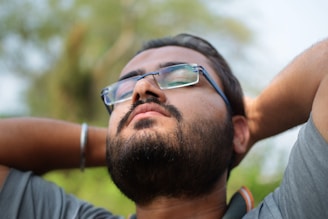 A person with facial hair and glasses appears relaxed, with closed eyes and hands resting behind the head. The background is softly blurred, indicating an outdoor setting with greenery.