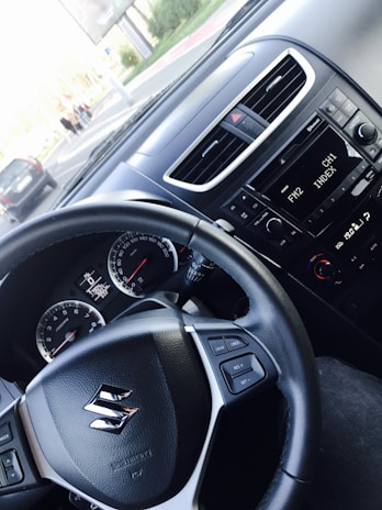 The interior view of a car focusing on the driver's area. The steering wheel, featuring a Suzuki logo and several control buttons, is prominent. The dashboard shows an instrument cluster with a speedometer and a digital display, while the car radio displays FM2 CH1. Outside the car, part of the road with a couple of vehicles and pedestrians is visible.