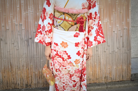 A person wearing a traditional Japanese kimono with a floral pattern featuring red and white flowers. The obi belt is intricately designed with golden and black patterns, accessorized with a red tassel and floral embellishment. The person is holding a matching handbag with similar floral designs. The background consists of a bamboo-like paneling, complementing the traditional theme.
