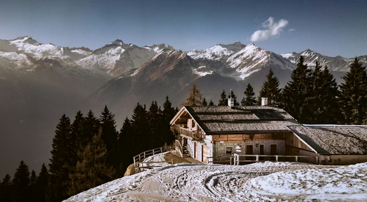 A timber and stone mountain refuge in the snow among conifers with a snow-covered mountain range behind, Madonna di Campiglio