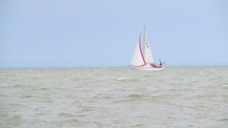 A sailboat with white sails and a red stripe is navigating through a body of water with gentle waves under a clear, light blue sky. There are two people on the boat, one standing near the mast and another near the stern.