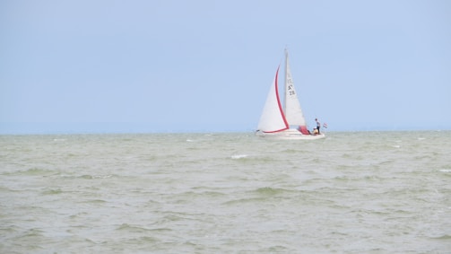 A sailboat with white sails and a red stripe is navigating through a body of water with gentle waves under a clear, light blue sky. There are two people on the boat, one standing near the mast and another near the stern.