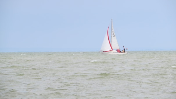 A sailboat with white sails and a red stripe is navigating through a body of water with gentle waves under a clear, light blue sky. There are two people on the boat, one standing near the mast and another near the stern.