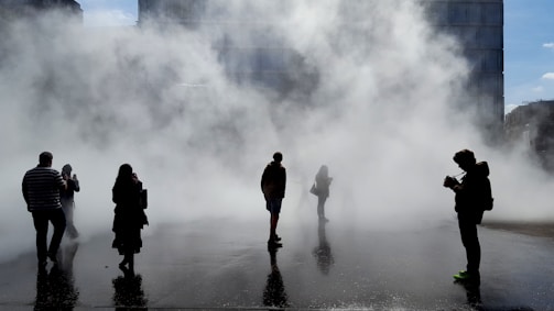 Blurred silhouettes of people walking through a misty urban street, suggesting hidden stories in plain sight.