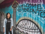 A woman stands smiling in front of a colorful mural with the words 'Welcome to Toronto' painted on it. The mural features iconic city landmarks including the CN Tower. The wall is composed of textured bricks painted in vibrant shades such as blue, purple, and black. The woman is wearing a black t-shirt with the words 'Home is North' and a light denim jacket.