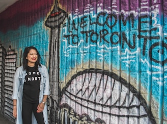 A woman stands smiling in front of a colorful mural with the words 'Welcome to Toronto' painted on it. The mural features iconic city landmarks including the CN Tower. The wall is composed of textured bricks painted in vibrant shades such as blue, purple, and black. The woman is wearing a black t-shirt with the words 'Home is North' and a light denim jacket.