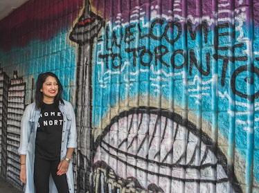 A woman stands smiling in front of a colorful mural with the words 'Welcome to Toronto' painted on it. The mural features iconic city landmarks including the CN Tower. The wall is composed of textured bricks painted in vibrant shades such as blue, purple, and black. The woman is wearing a black t-shirt with the words 'Home is North' and a light denim jacket.