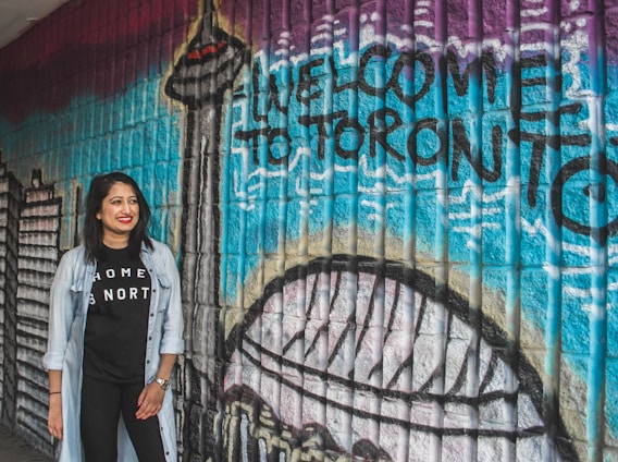 A woman stands smiling in front of a colorful mural with the words 'Welcome to Toronto' painted on it. The mural features iconic city landmarks including the CN Tower. The wall is composed of textured bricks painted in vibrant shades such as blue, purple, and black. The woman is wearing a black t-shirt with the words 'Home is North' and a light denim jacket.