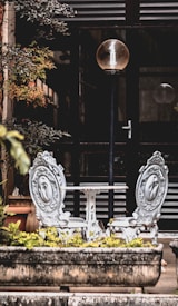 Two ornate, vintage-style chairs with intricate designs face each other across a small round table. A tall street lamp with a round bulb is positioned between them. Lush greenery surrounds the seating area, and there is a potted plant to the left. The background consists of a building entrance with a glass door and horizontal slats.