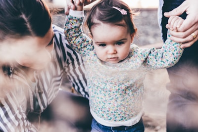 A toddler in a floral-patterned top is being supported by adults holding her hands as she takes steps. One adult is crouched beside her, wearing a striped shirt. The scene conveys a familial and nurturing atmosphere.