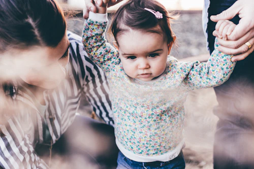 A toddler in a floral-patterned top is being supported by adults holding her hands as she takes steps. One adult is crouched beside her, wearing a striped shirt. The scene conveys a familial and nurturing atmosphere.