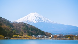 A picturesque view of a snow-capped mountain, likely Mount Fuji, dominates the background with a clear blue sky. The foreground features a serene lake surrounded by a vibrant green forest and small buildings. The scene is calm and visually stunning with natural beauty.
