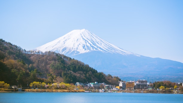 A picturesque view of a snow-capped mountain, likely Mount Fuji, dominates the background with a clear blue sky. The foreground features a serene lake surrounded by a vibrant green forest and small buildings. The scene is calm and visually stunning with natural beauty.
