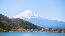 A picturesque view of a snow-capped mountain, likely Mount Fuji, dominates the background with a clear blue sky. The foreground features a serene lake surrounded by a vibrant green forest and small buildings. The scene is calm and visually stunning with natural beauty.