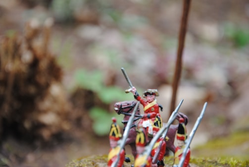 A miniature figure depicting a soldier on horseback raises a sword amidst a background of blurred foliage. The figure is detailed with a red and yellow uniform, complemented by a horse adorned in similar colors. Nearby, other figures with spears add to the scene of a miniature battle scenario.