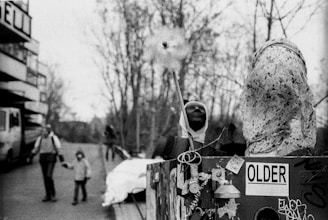 A black and white image with a focus on a textured, abstract statue placed on a decorated structure featuring various stickers and tags, including a notable 'OLDER' label. In the background, blurred silhouettes of people, including an adult holding a child's hand, walk along a street bordered by a building and leafless trees.