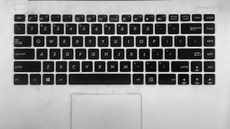 A monochrome image of a laptop keyboard featuring standard QWERTY layout. Keys include letters, numbers, and function keys as well as shortcuts such as Ctrl, Alt, and Windows key. The surface is slightly worn, indicating use. Below the keyboard, a touchpad is visible.